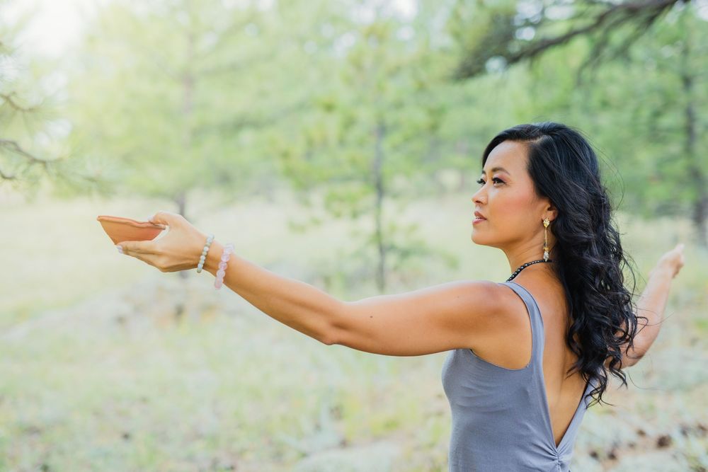 Woman practicing yoga outdoors in a forest, arms extended in a graceful pose wearing a gray tank top.