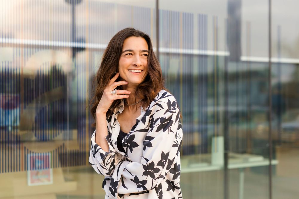 Someone in a black and white floral print top smiles while standing in front of a glass building exterior.