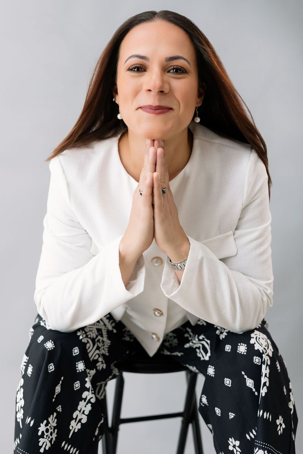 Woman in white blouse and patterned skirt sitting with hands pressed together in prayer gesture, smiling.