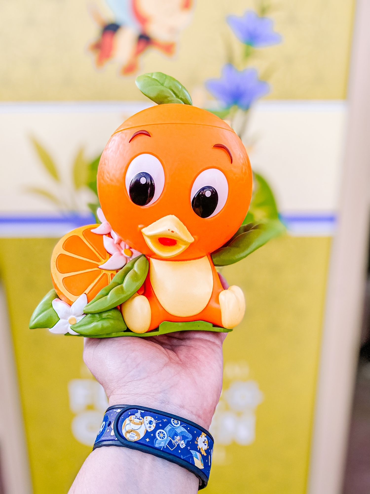 Cute orange plush toy mascot character with green leaves sits atop a hand wearing a blue patterned bracelet.
