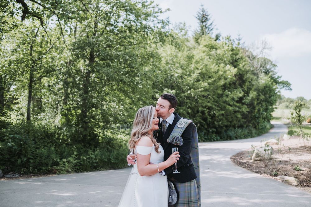 Newlywed couple share a kiss outdoors with lush green trees behind them.
