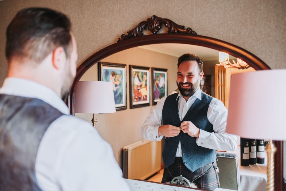 Smiling groom in kilt and waistcoat adjusts outfit while looking in mirror.