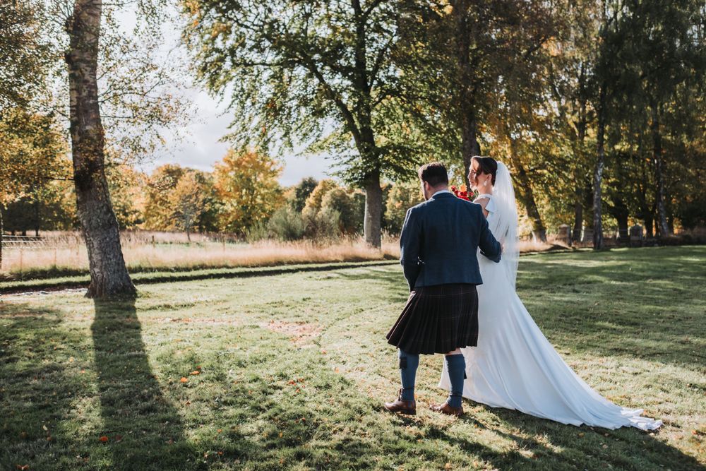 Bride and groom walk hand in hand through sunny autumn woodland grounds.