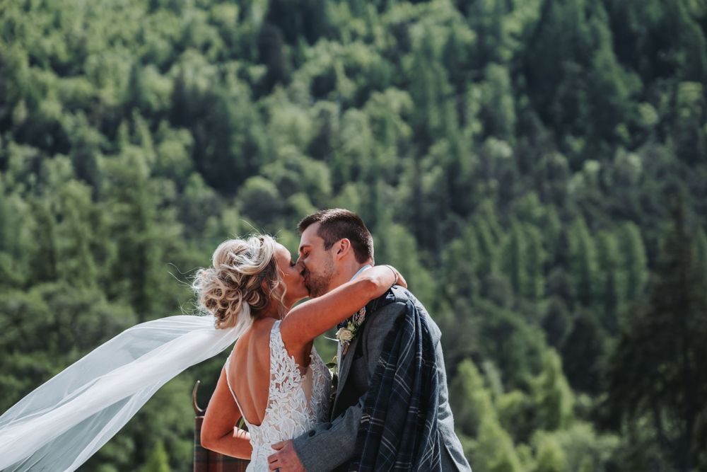 Bride and groom kissing with wedding veil blowing in the wind against a lush forest.