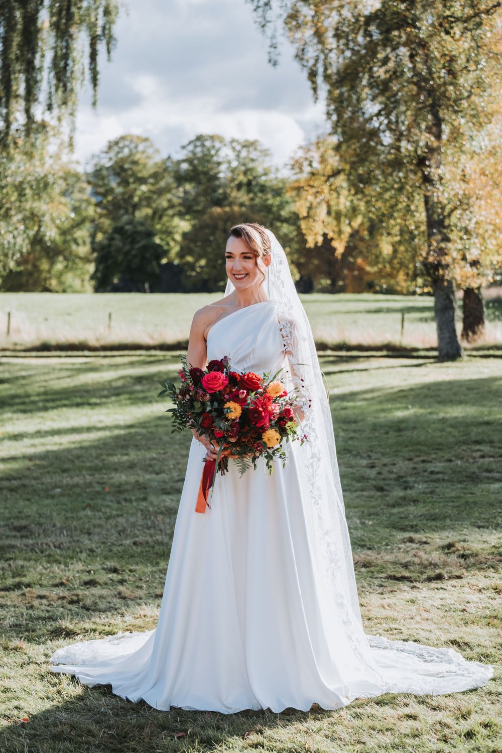 Bride in white one-shoulder gown holds colorful bouquet in sunny garden setting.
