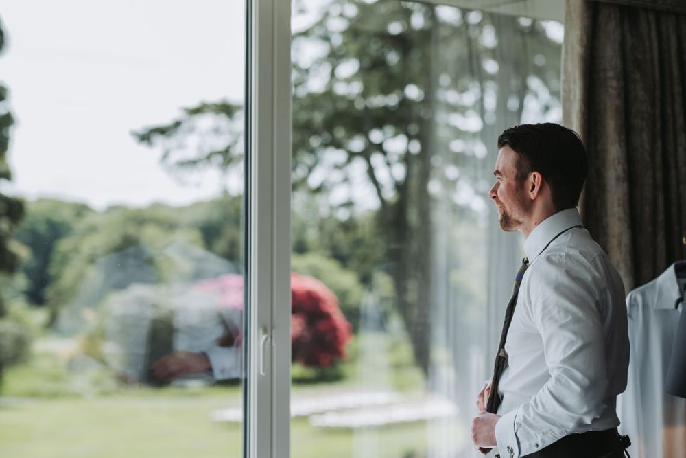 Groom in white shirt and suspenders gazing out large window at lush green garden before wedding ceremony.