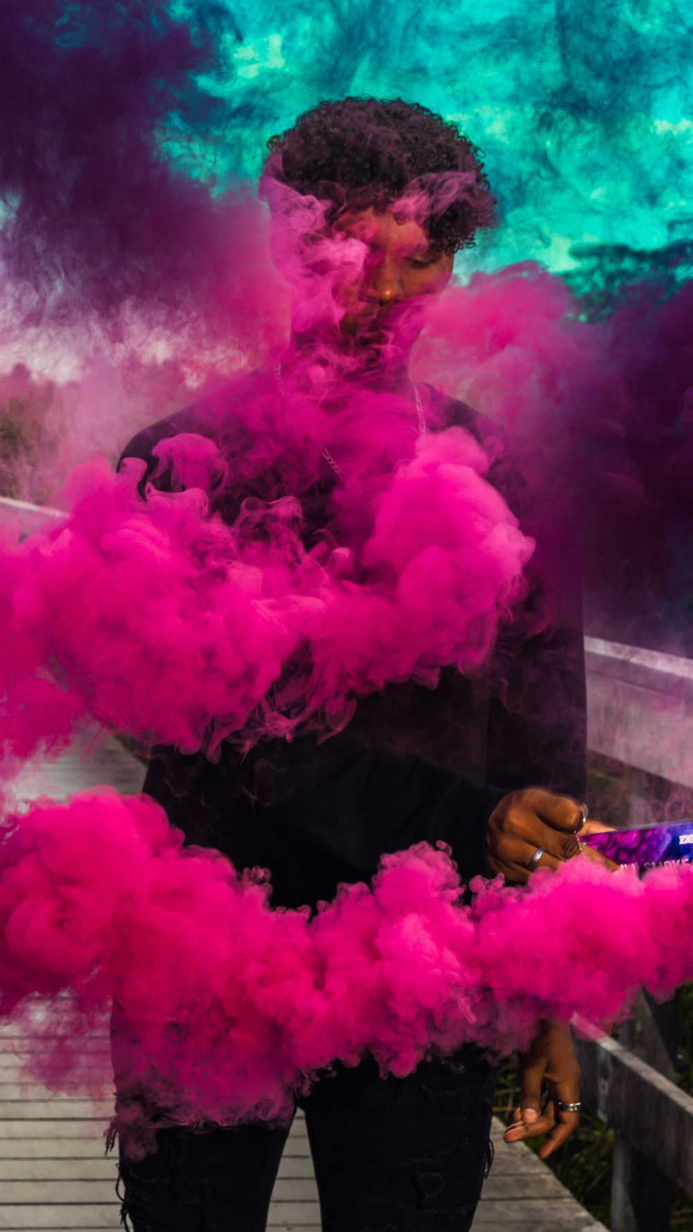 Figure surrounded by vibrant pink smoke and turquoise clouds creates dramatic artistic portrait with colored smoke bombs.