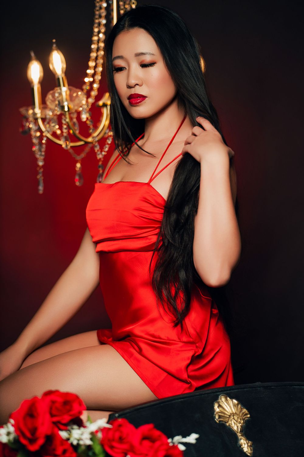 Elegant red satin dress photographed with roses and gold chandelier against dark moody background.