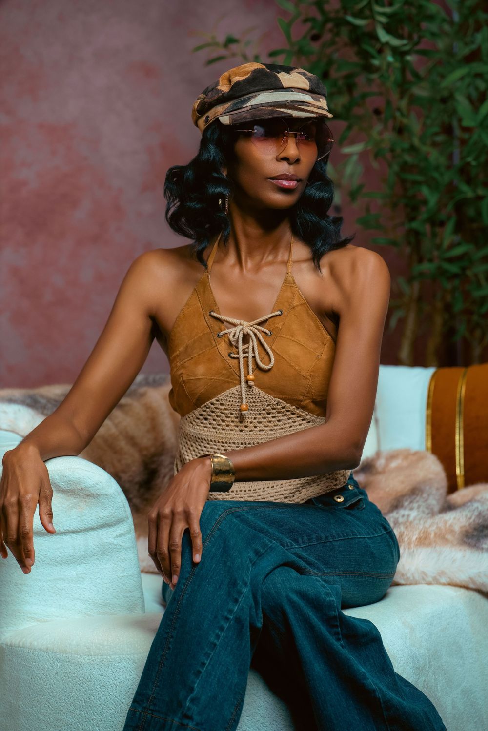 A woman in a crochet halter top, jeans, and camo hat sits confidently on a white couch in a stylish studio setting.