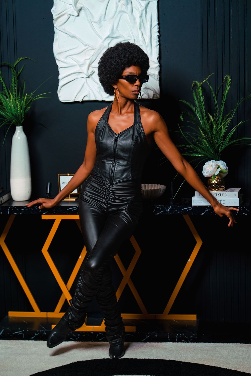 A stylish woman in a black leather jumpsuit and sunglasses poses confidently in a chic, dark-themed interior.