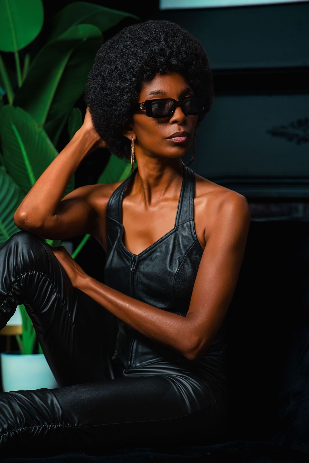 A stylish Black woman with an afro hairstyle wearing black sunglasses, leather halter top, and gloves poses confidently indoors.