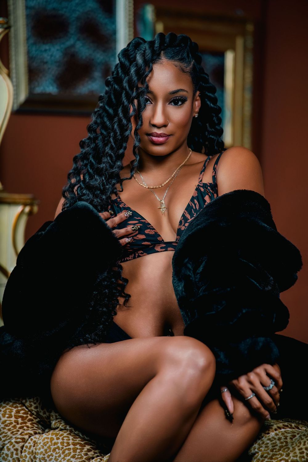 A woman with long curly braids wearing a black lace bralette and velvet coat poses on a leopard print chair.