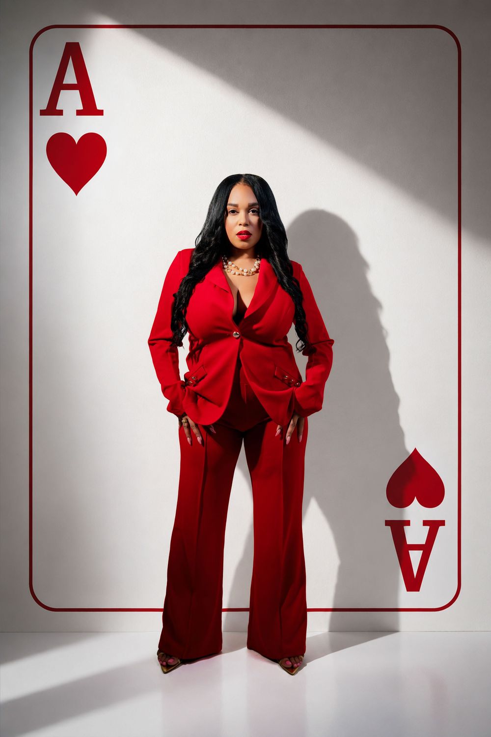 Woman in red suit poses confidently against white wall with Ace of Hearts playing card design backdrop.