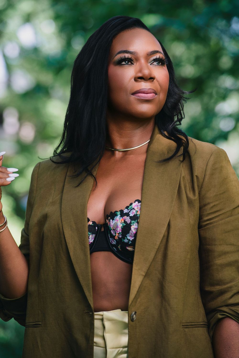 Woman in olive blazer and floral bralette poses outdoors with lush green trees in background.