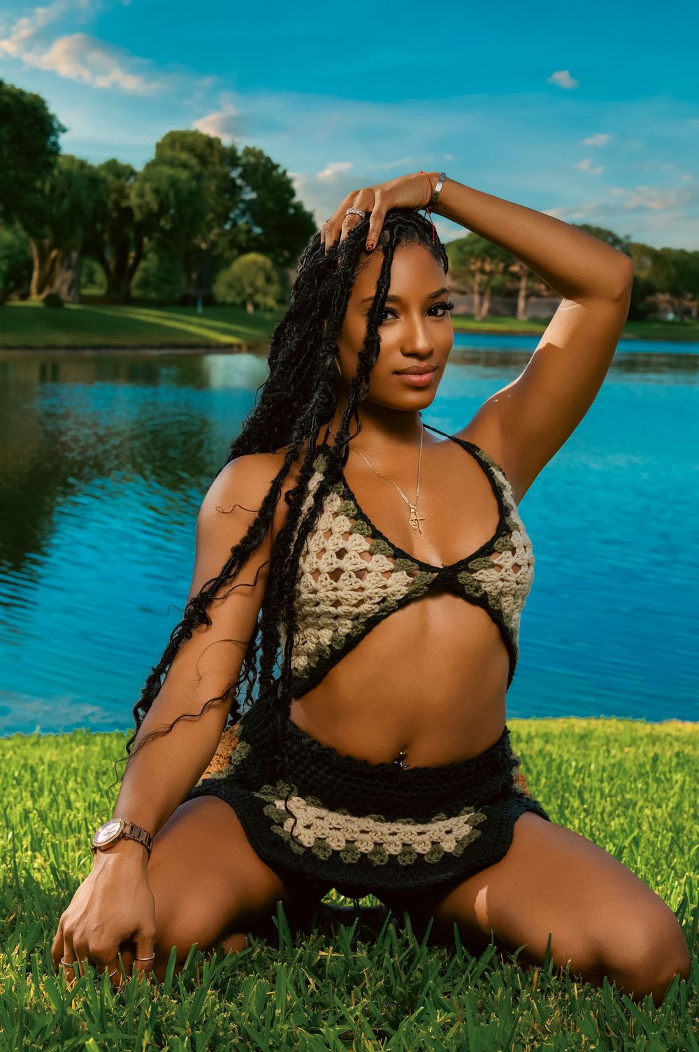 A model poses in black lace lingerie next to a scenic lake on a sunny day with green grass and blue water.