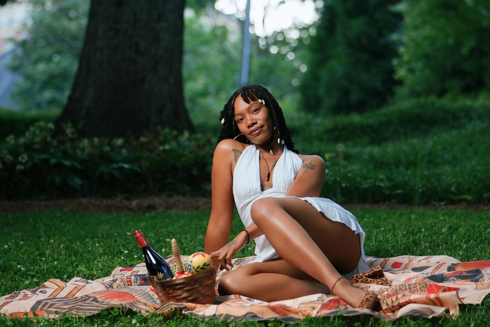 Woman in white dress enjoying a picnic on a blanket in a lush green park with wine and fruit.