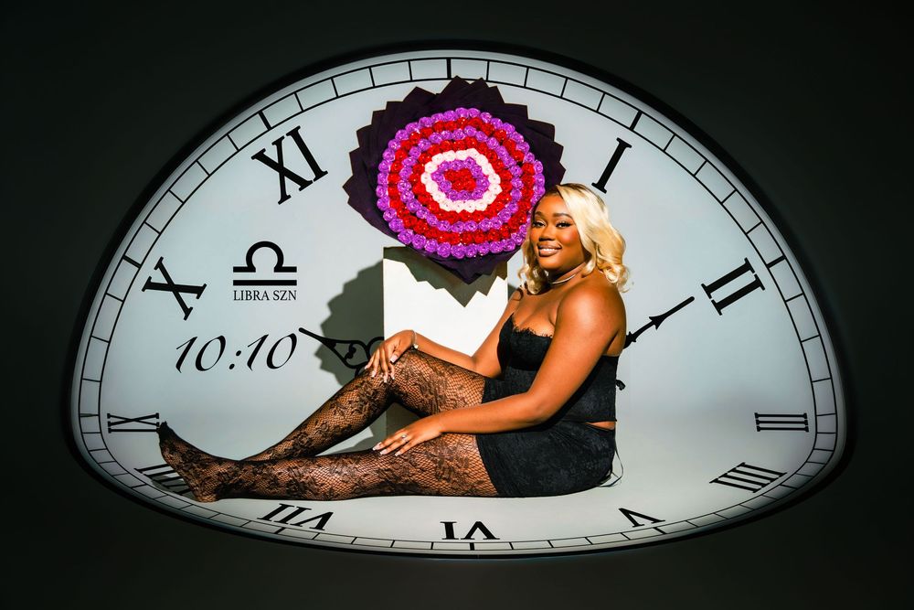 Woman posing in front of a half-clock backdrop showing 10:10, with a colorful floral mandala and Libra zodiac sign.