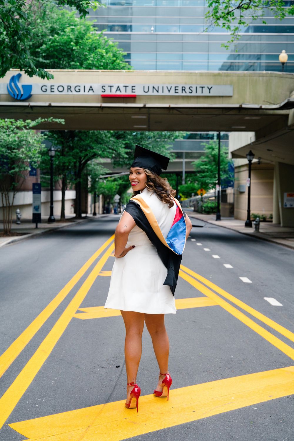 Georgia State University graduate in cap and gown poses on yellow crosswalk lines in white dress and red heels.