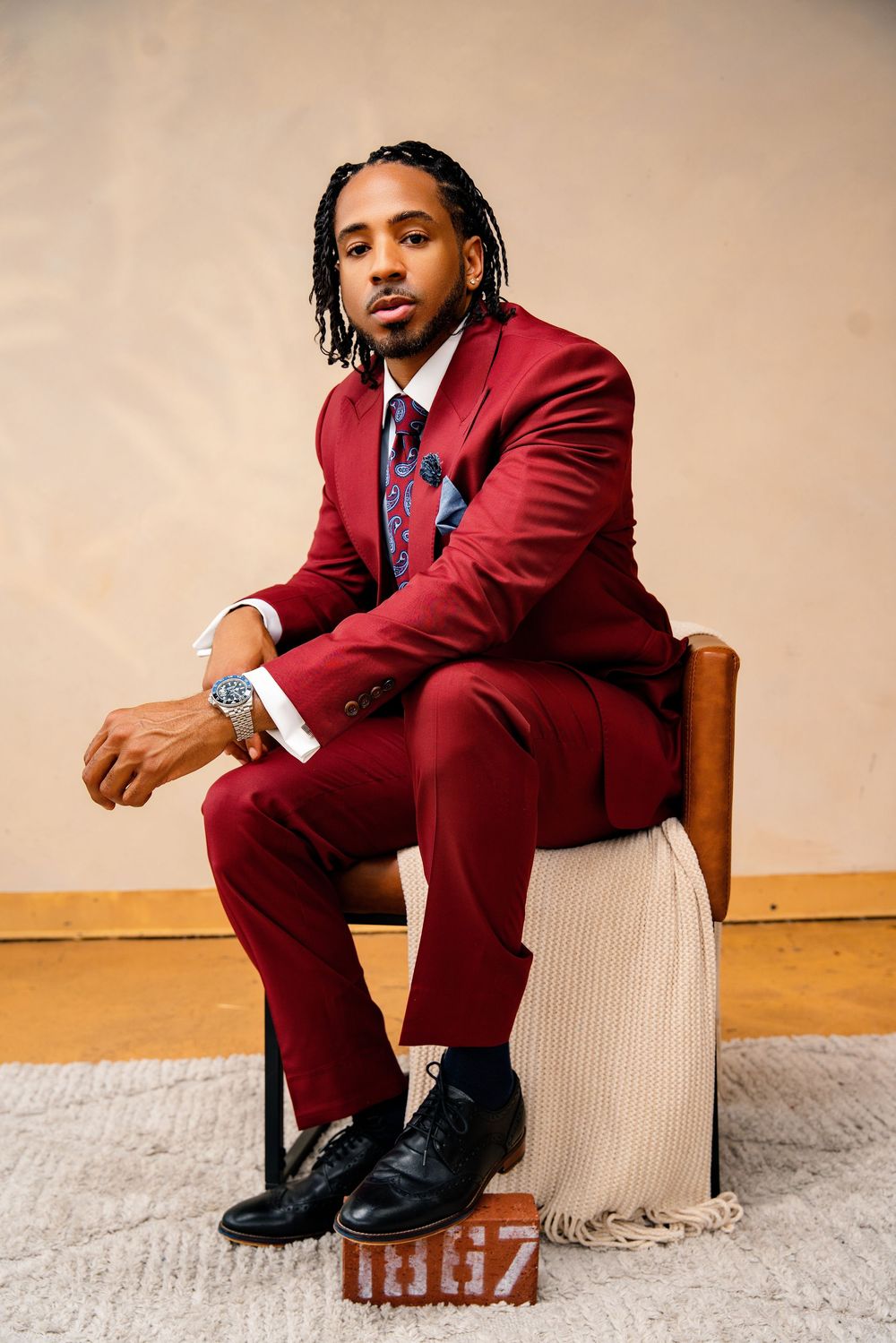 Man in burgundy suit with braids sits confidently on chair, wearing patterned tie and dress shoes.