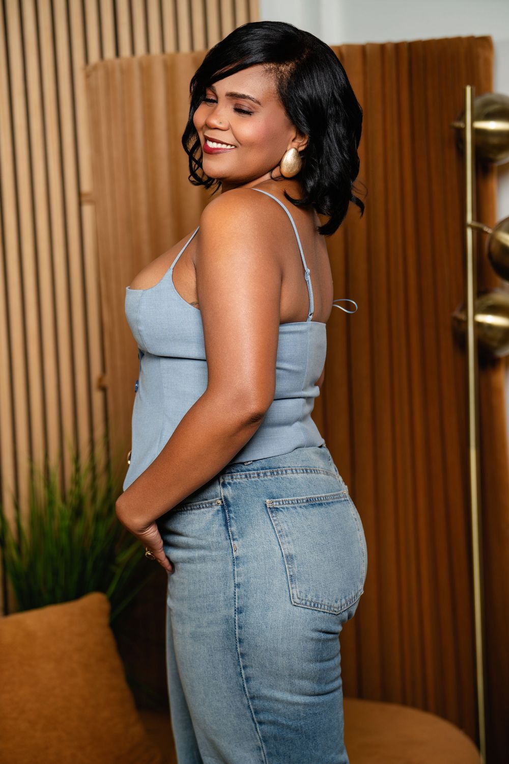 Woman wearing light blue tank top and jeans poses confidently against wooden paneled wall in casual photoshoot.
