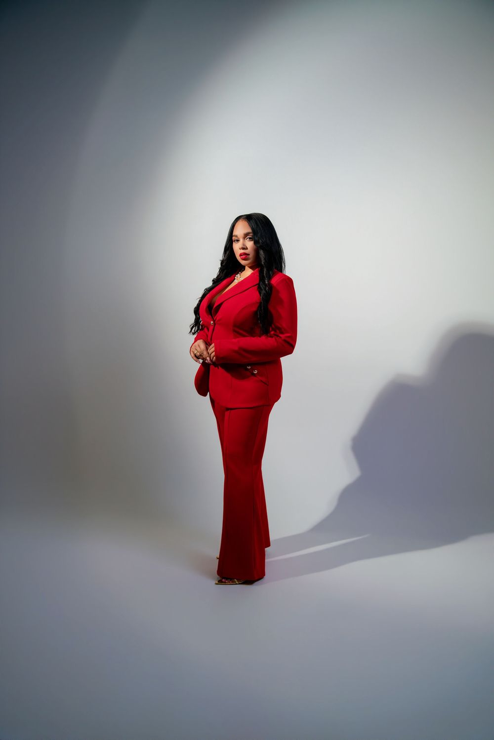 Woman in a red blazer and flared pants suit poses confidently in a studio with dramatic lighting and shadow.