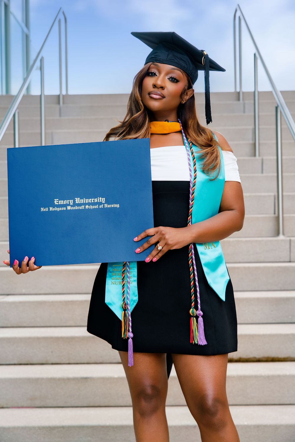 A proud graduate holds her Emory University Nell Hodgson Woodruff School of Nursing diploma wearing cap and colorful stoles.