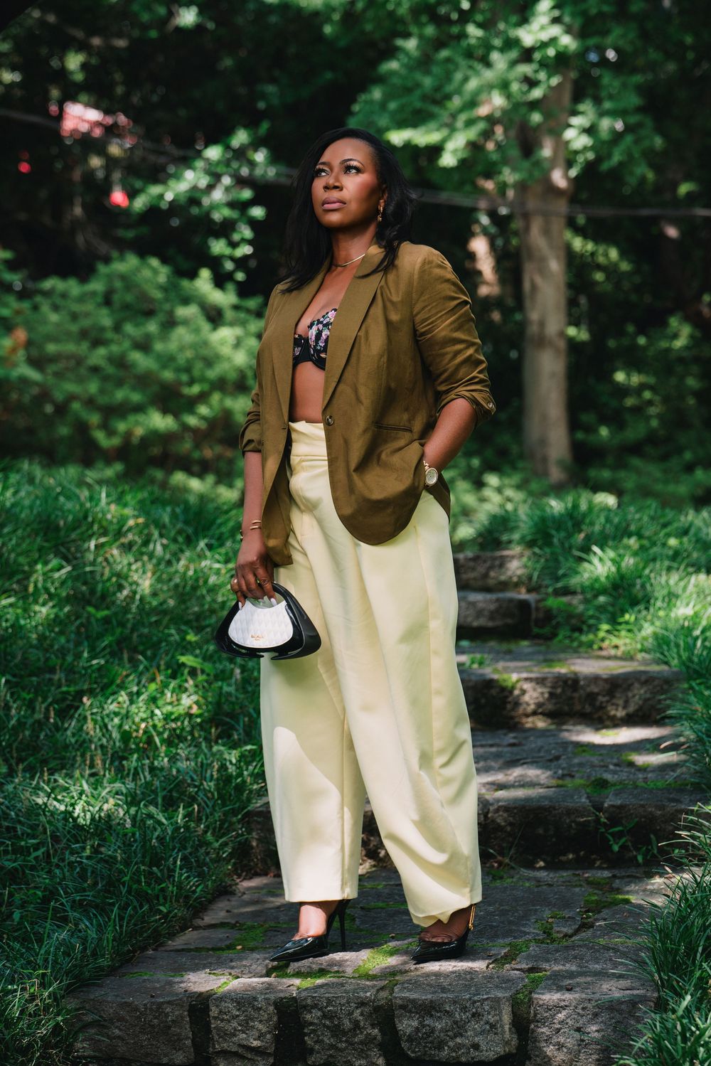 Woman in olive blazer, cream wide-leg pants, holding clutch, standing on stone steps surrounded by lush greenery.