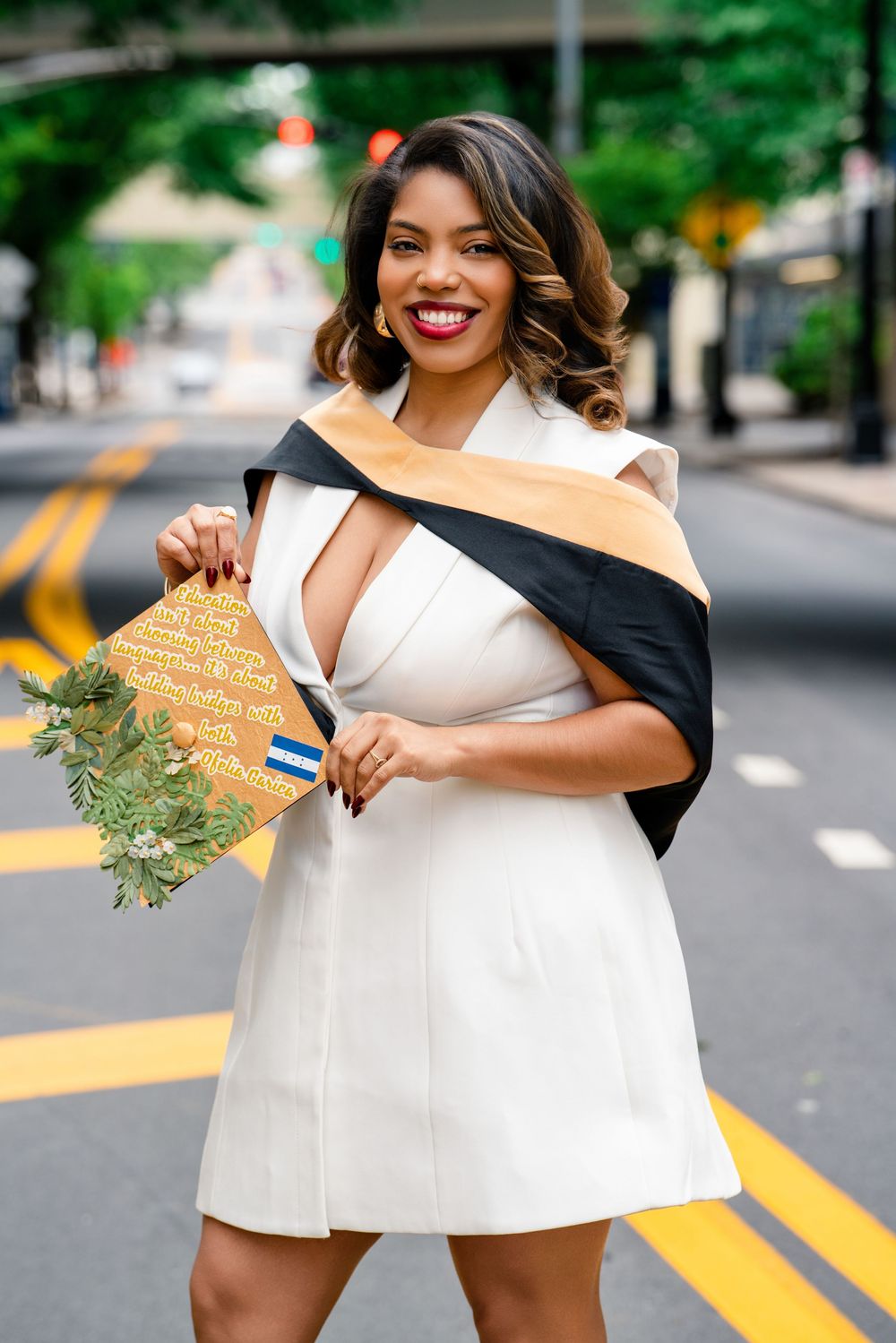 Smiling graduate in white dress holding decorated cap and flowers, wearing academic stole on city street.