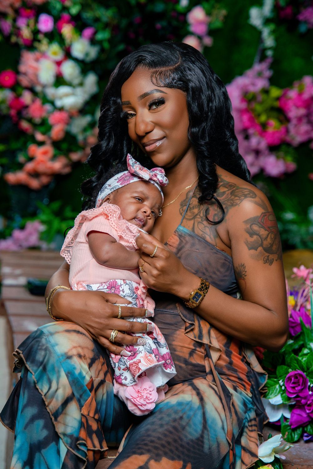 A smiling woman with tattoos holds a newborn baby in pink floral outfit, posed in front of colorful flower backdrop.