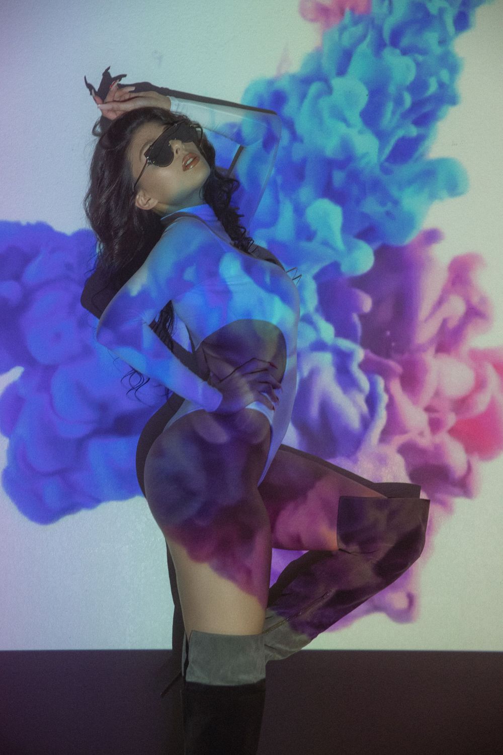 Artistic portrait with vibrant blue and purple smoke effects surrounding a figure wearing sunglasses and twirling in a dress.