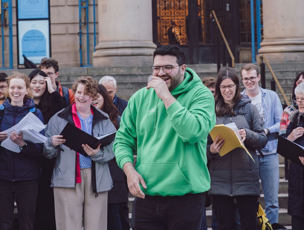 A person in a bright green hoodie stands smiling in front of a group of people holding papers outside a building.