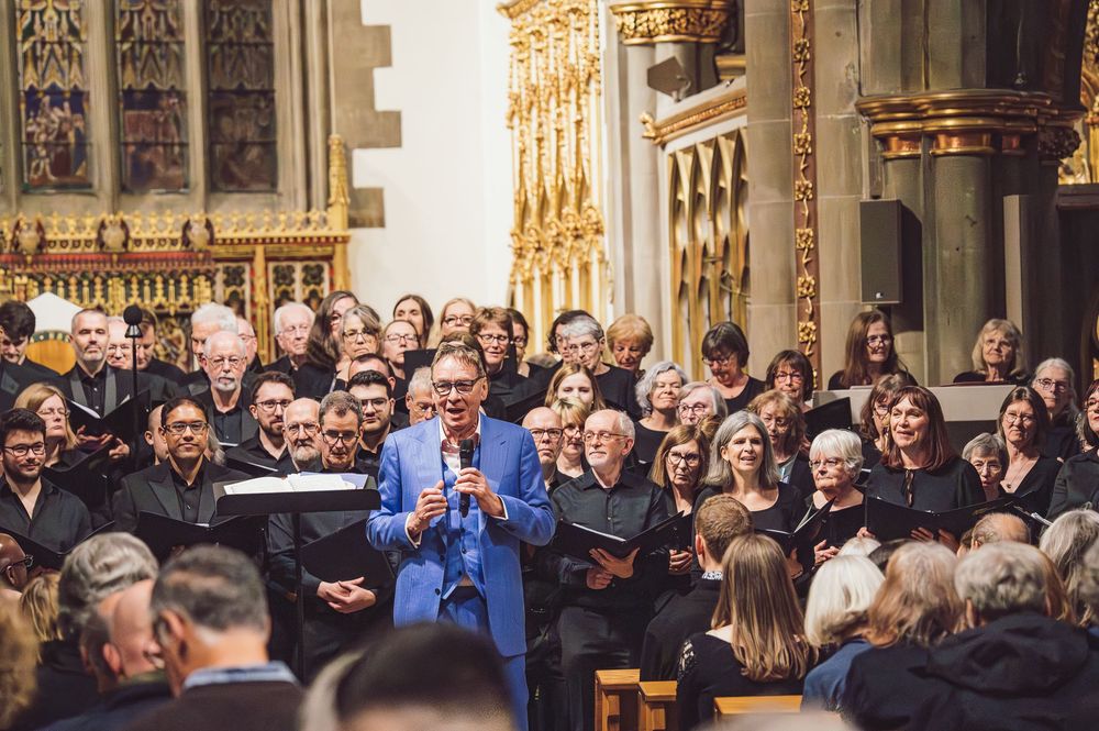 A large choir performs together during a concert inside a grand cathedral with ornate architecture and stained glass windows.