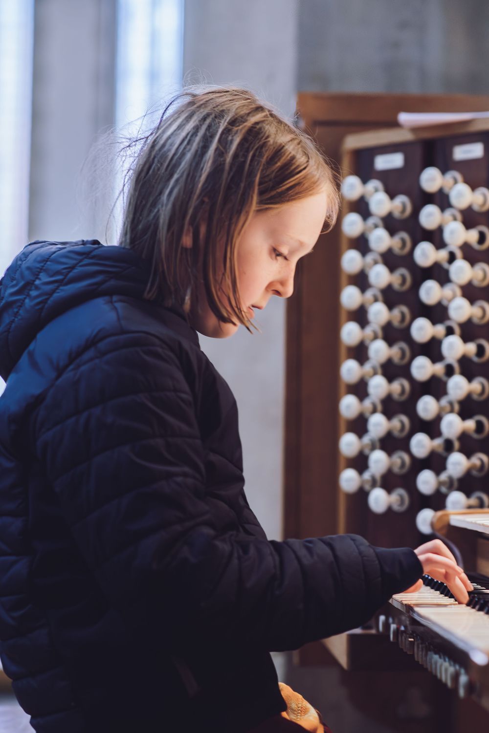 Young musician in a navy coat plays a church organ with many white stops visible in the background.