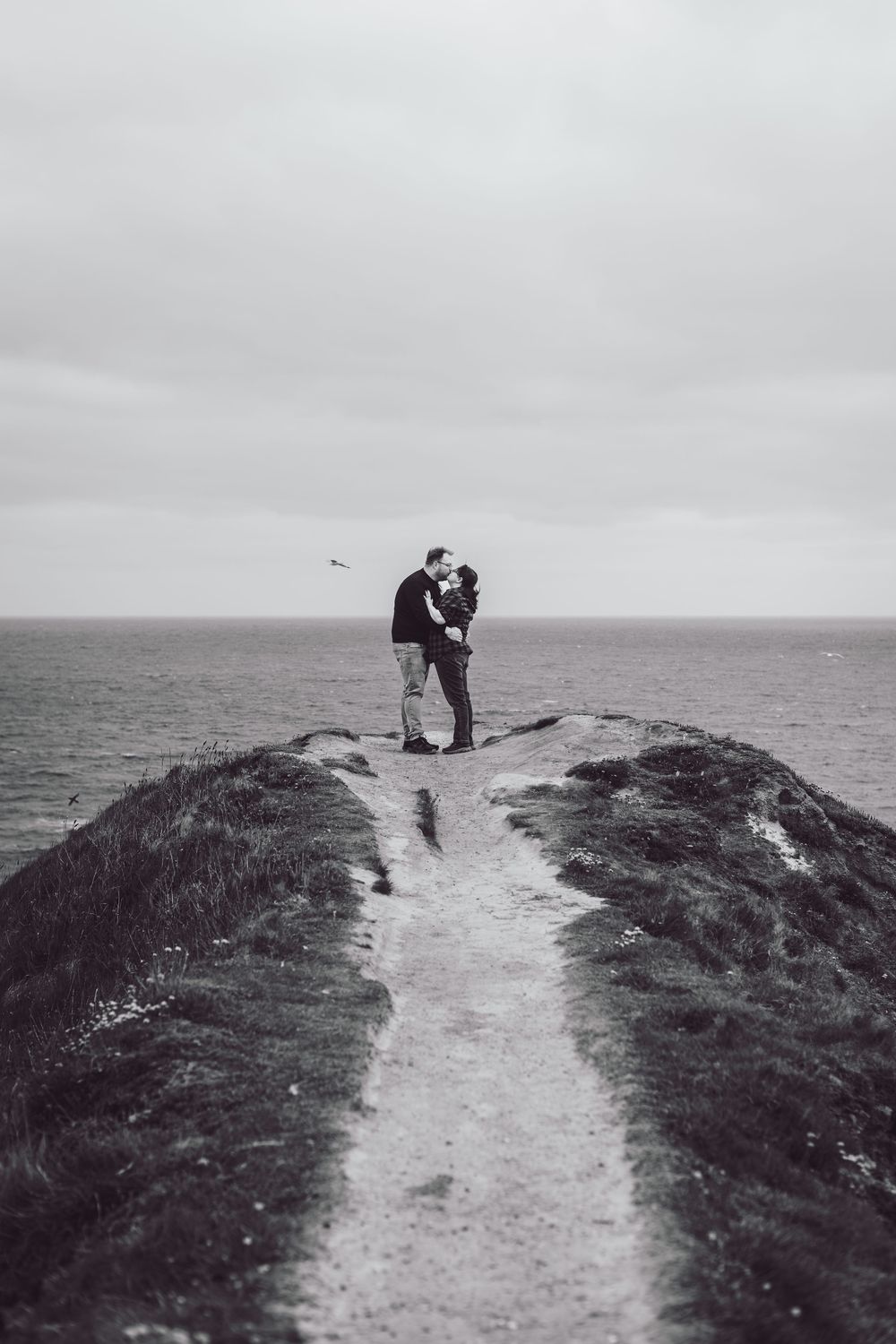 Two silhouettes embrace on a scenic coastal cliff path against moody gray skies and ocean horizon in black and white.