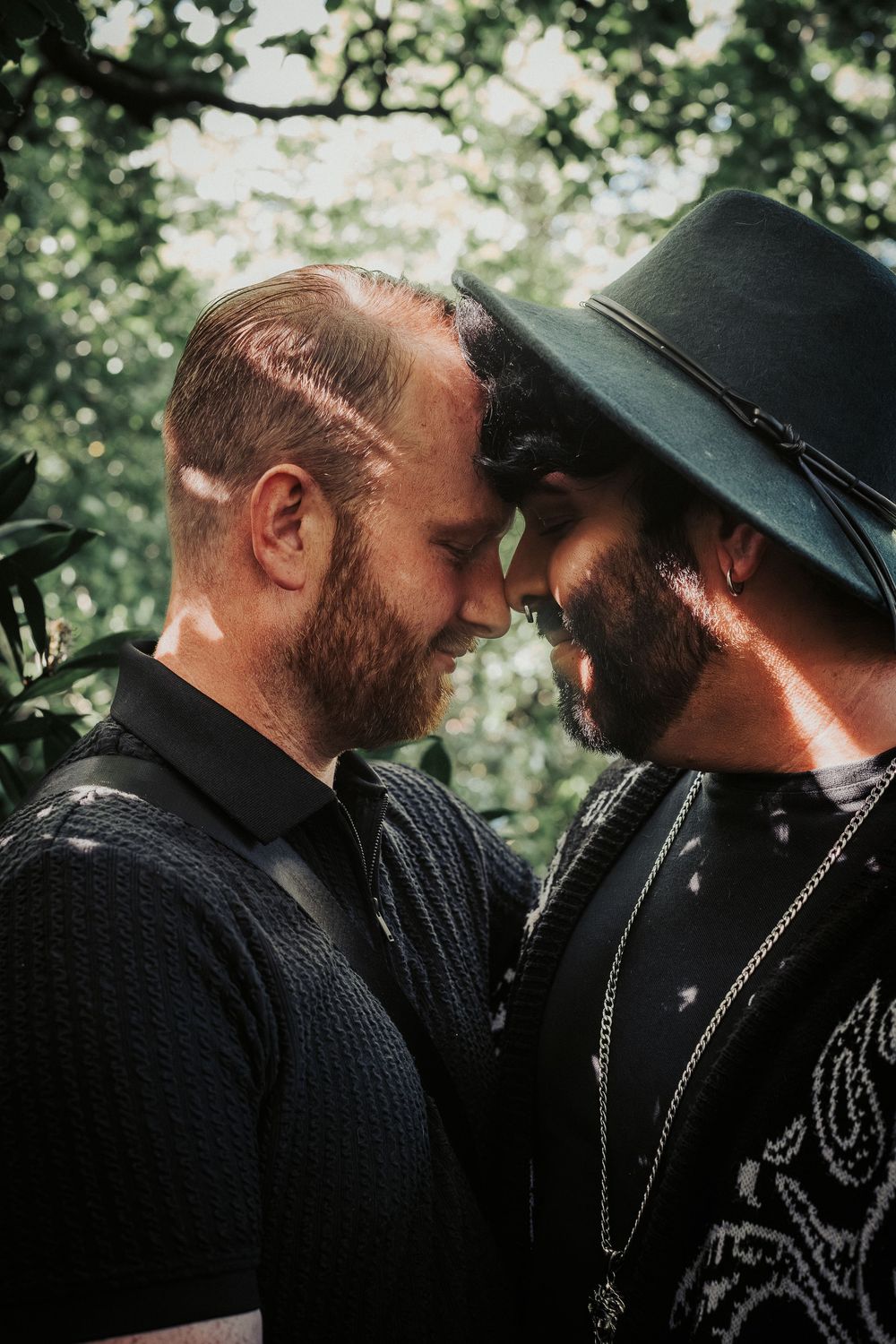 Two men in black clothing share an intimate moment in a lush green garden setting.