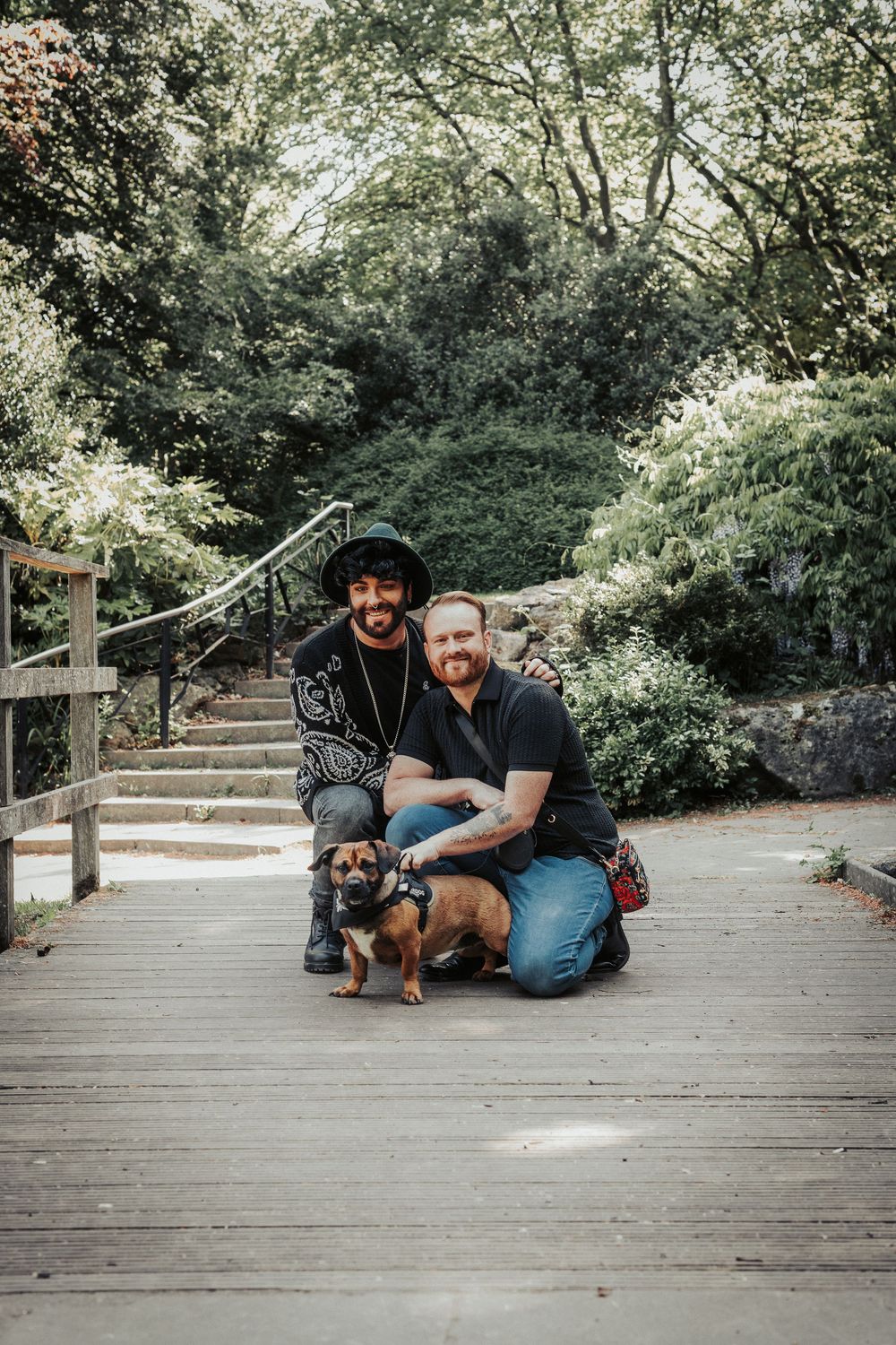 Two people kneeling on a wooden deck with their small dog amidst lush garden greenery and stone steps.