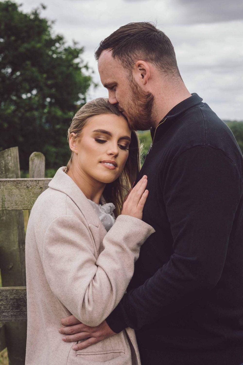 A romantic couple embraces outdoors against a rustic wooden fence on an overcast day.