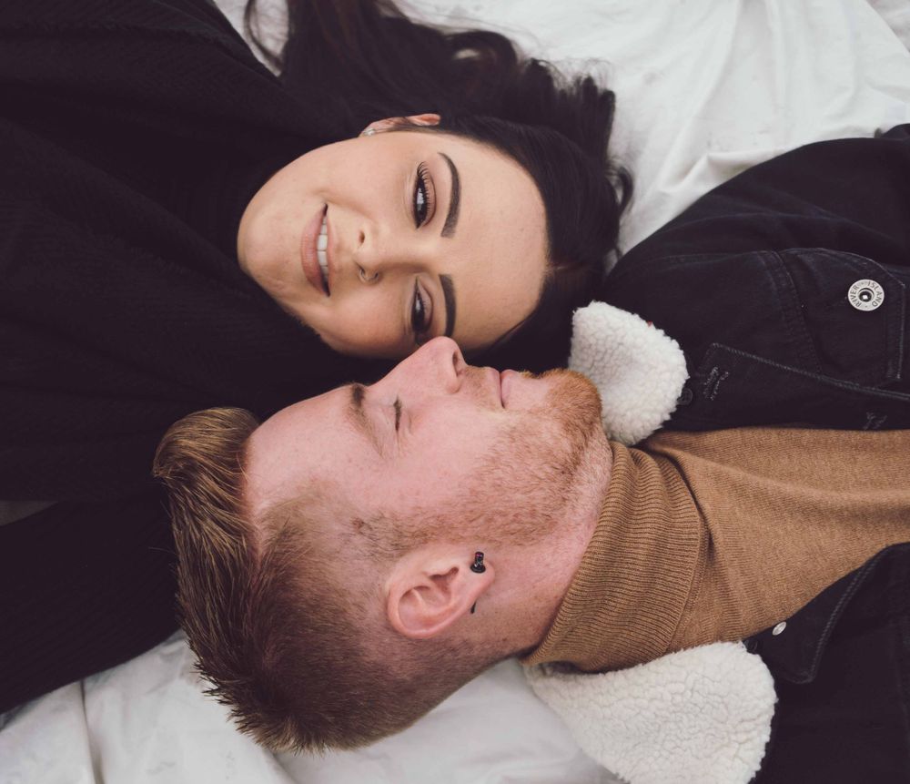 A couple lies on white bedding while wearing sweaters, with their heads close together in an aerial view portrait.