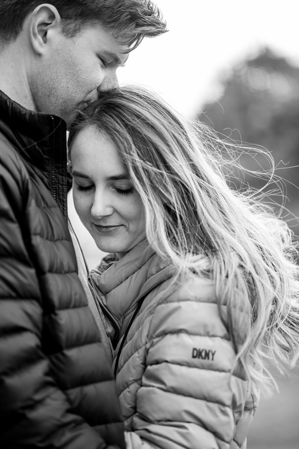 A romantic black and white portrait of a couple in puffy winter jackets embracing as wind blows through her hair.