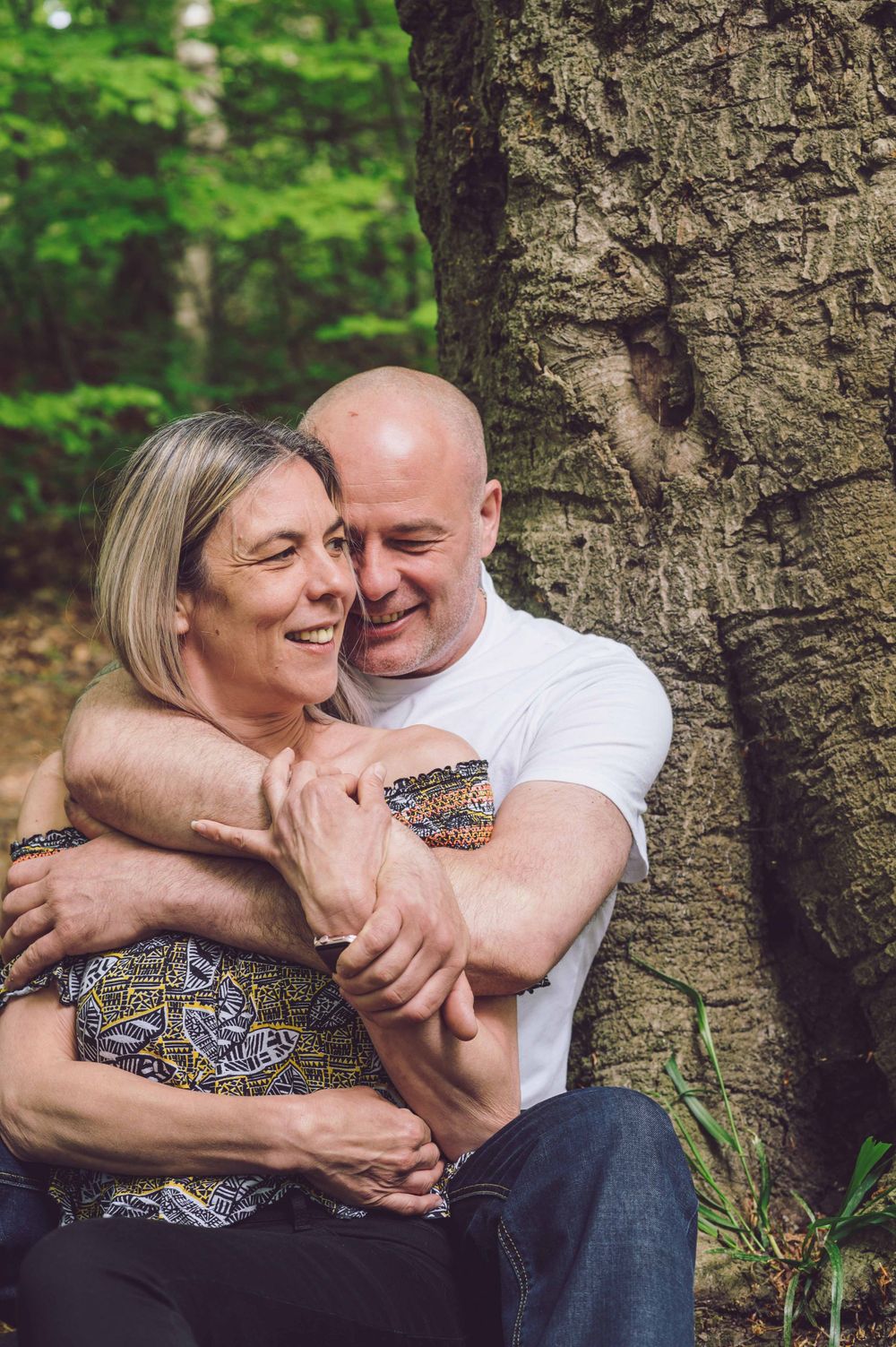 A couple shares an intimate embrace while sitting against a large tree trunk in a lush forest setting.