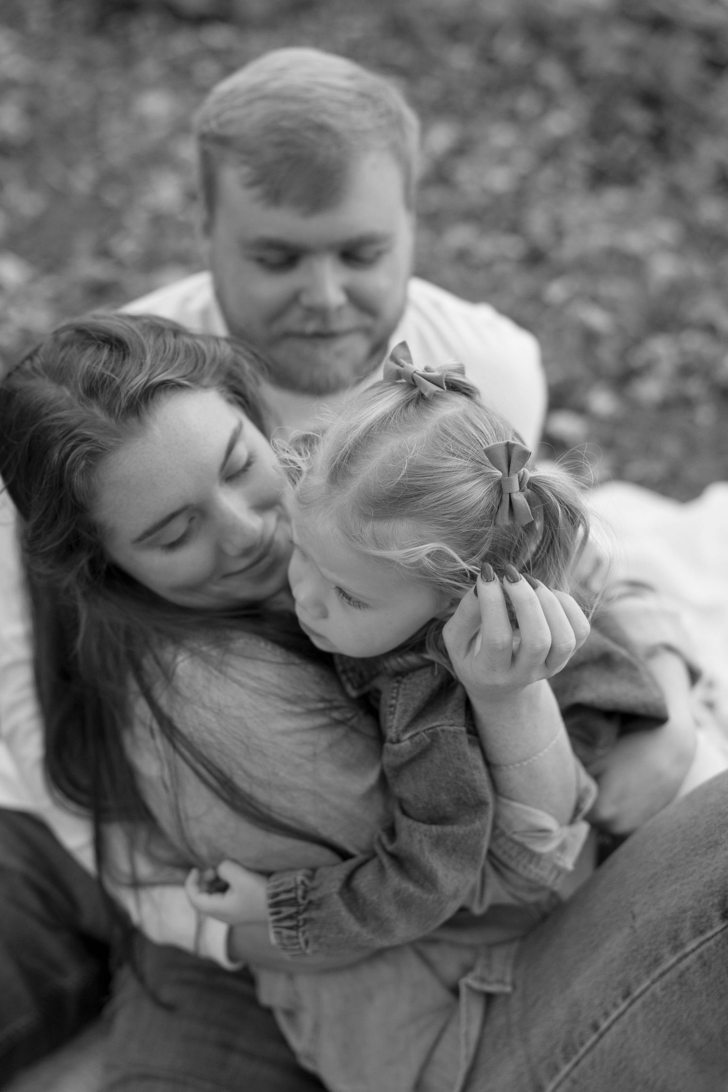 A family of three shares an intimate black and white moment outdoors while cuddling together on the ground.
