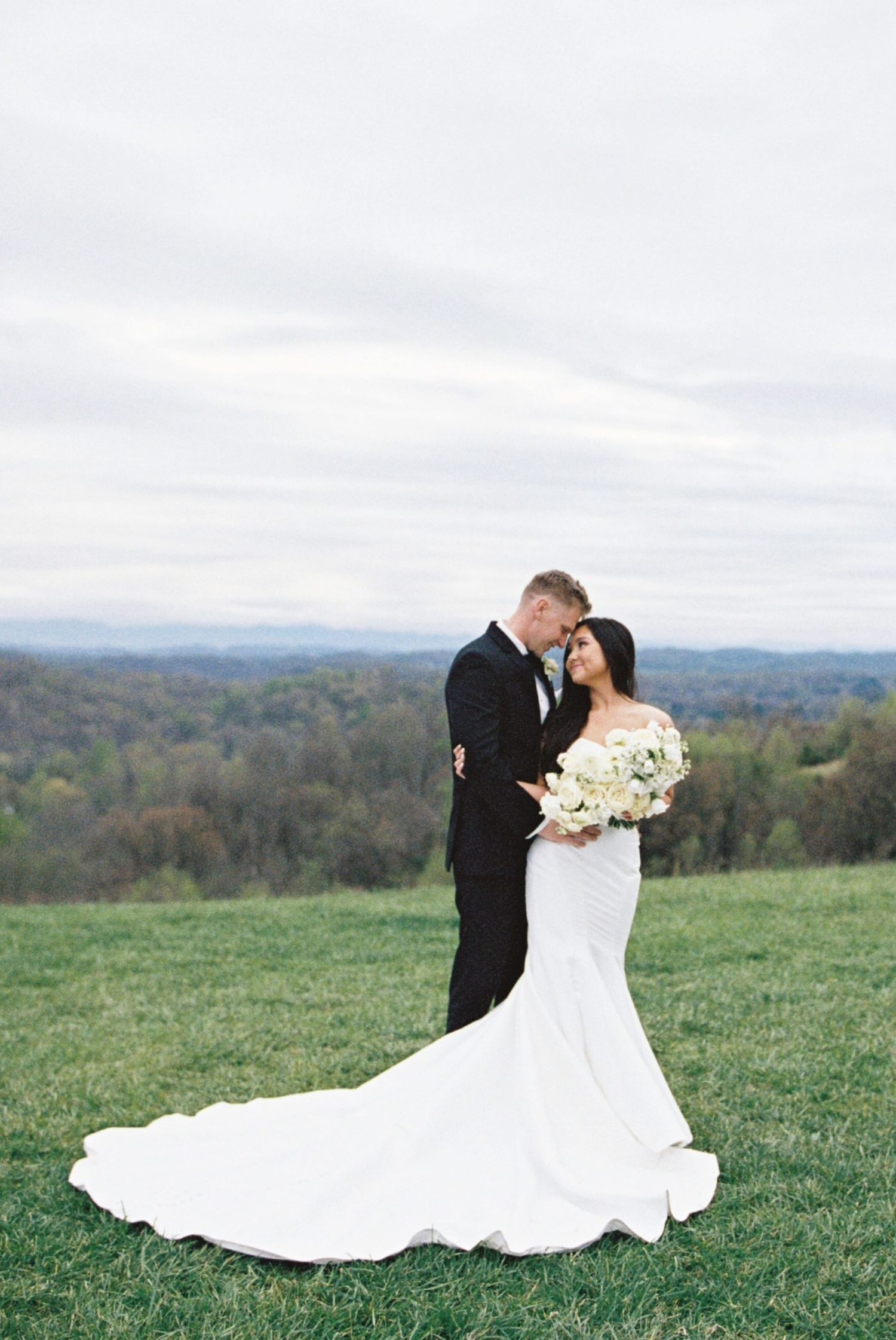 A couple shares a romantic moment on a hilltop overlooking an spring valley landscape while holding a white bouquet.