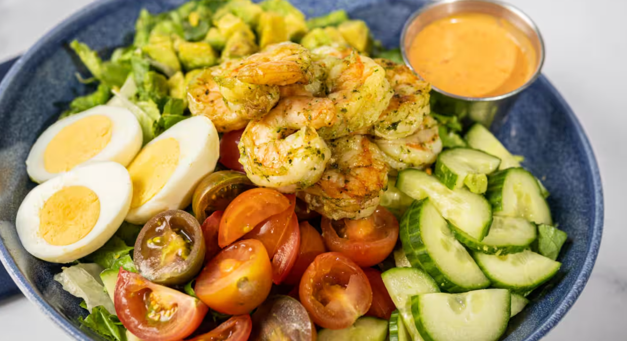 Fresh shrimp salad bowl with boiled eggs, tomatoes, cucumbers, lettuce and spicy orange aioli sauce on blue plate.