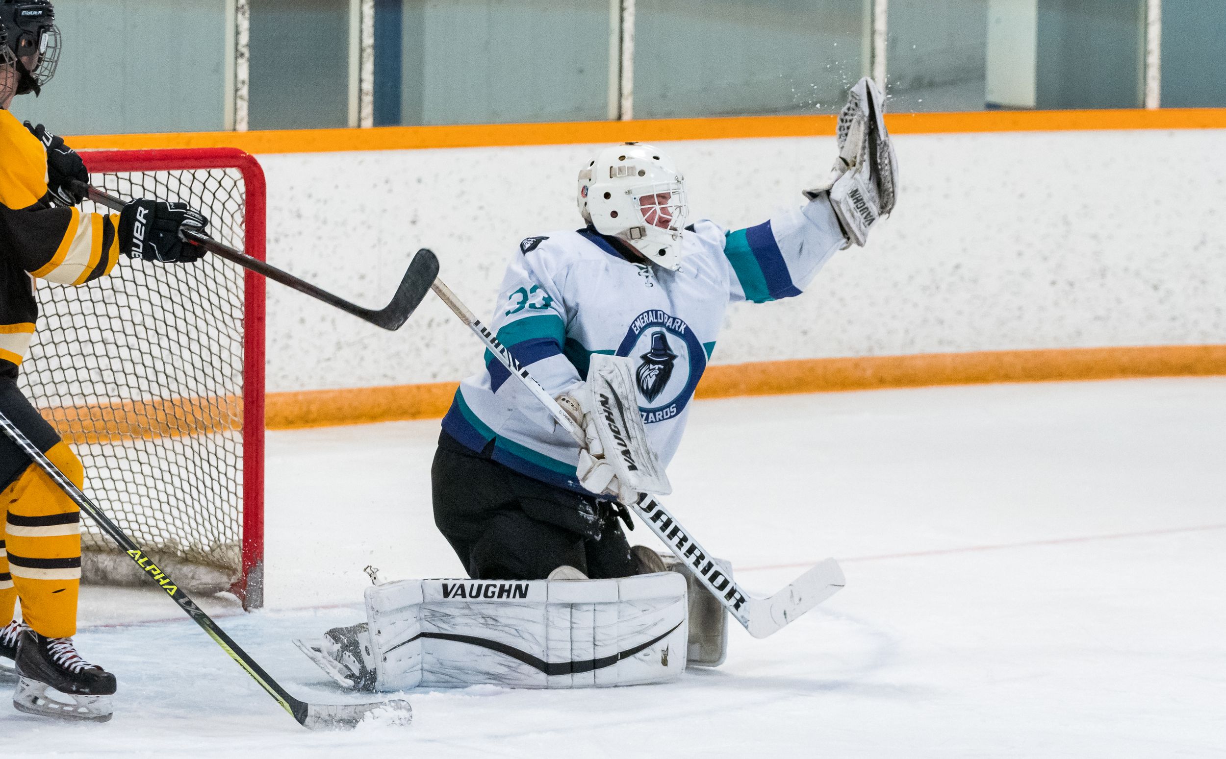 Ice hockey goalie in teal and black jersey makes a glove save during game against team in yellow uniforms.
