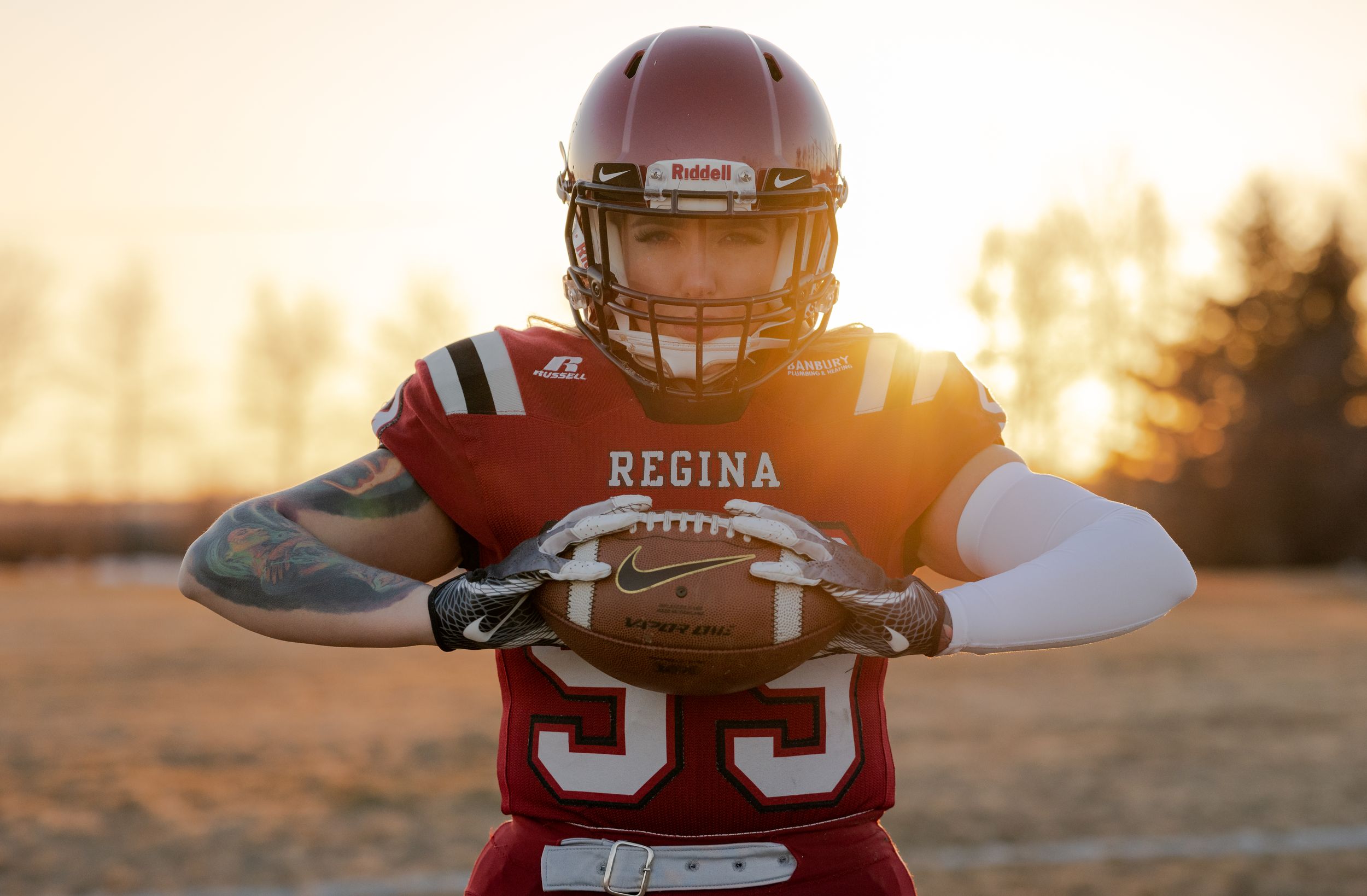 Football player in red Regina jersey holding ball at sunset during golden hour on field.