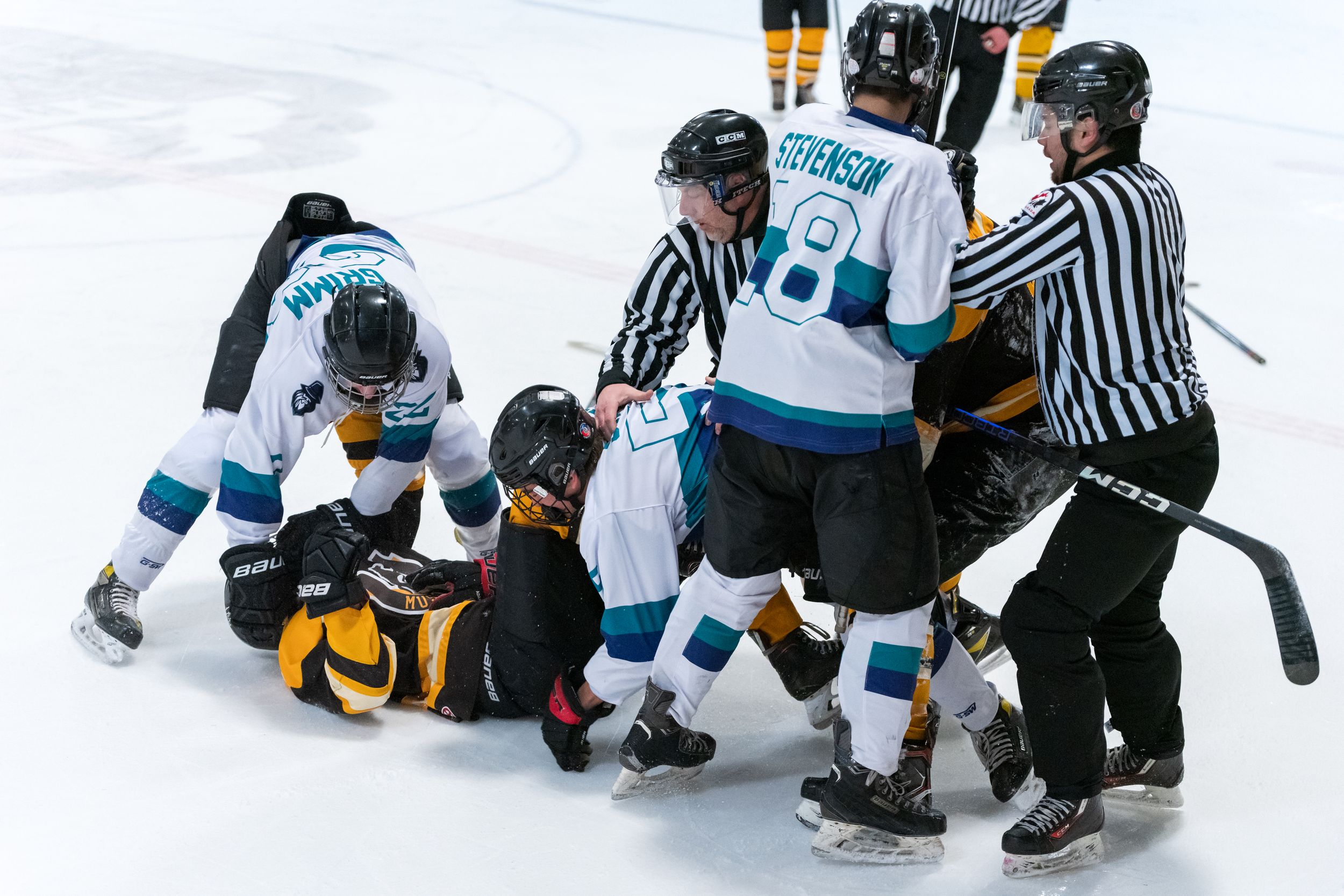Hockey players and referees gather around a fallen player during an on-ice altercation in a game between teams in white and black.