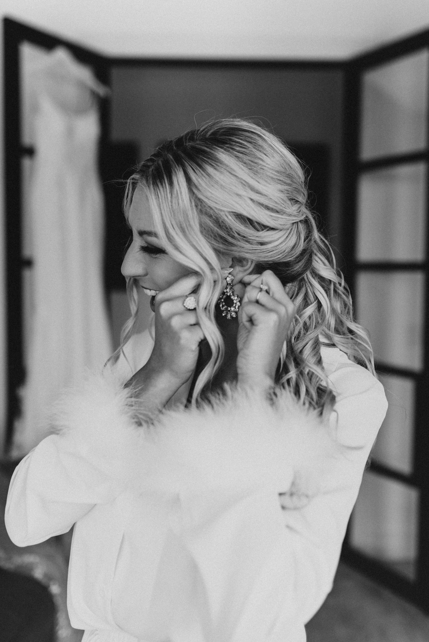 A bride getting ready in a black and white photo puts on earrings while wearing a delicate white top.