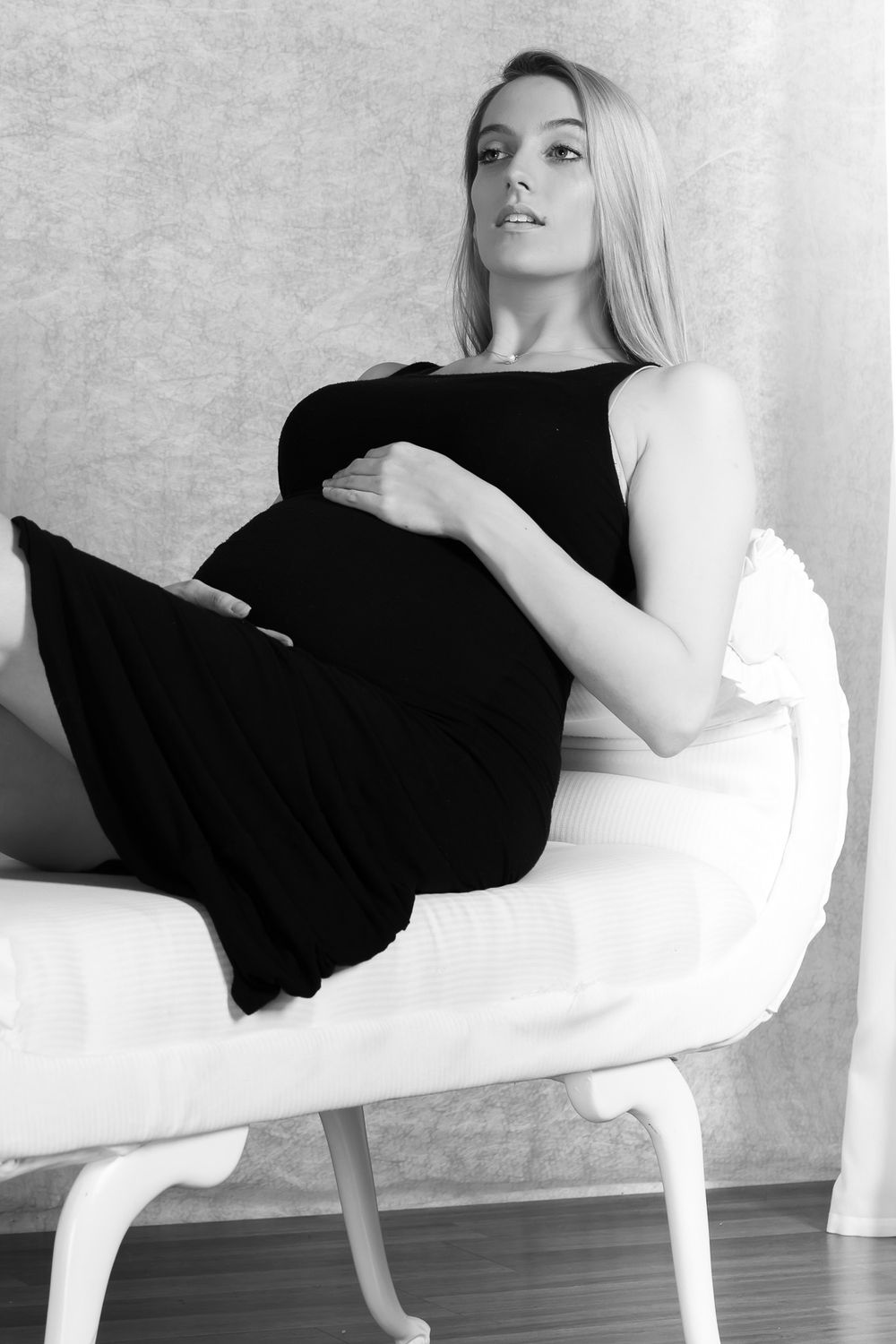 Maternity session and black and white portrait of someone wearing a black dress while reclining elegantly on a white modern chair.