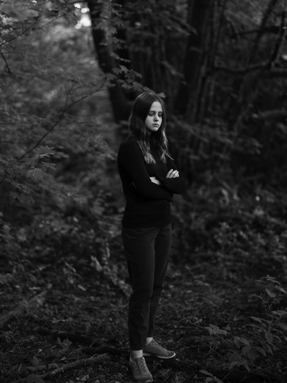 Dramatic black and white portrait of someone standing in a dark forest with arms crossed.