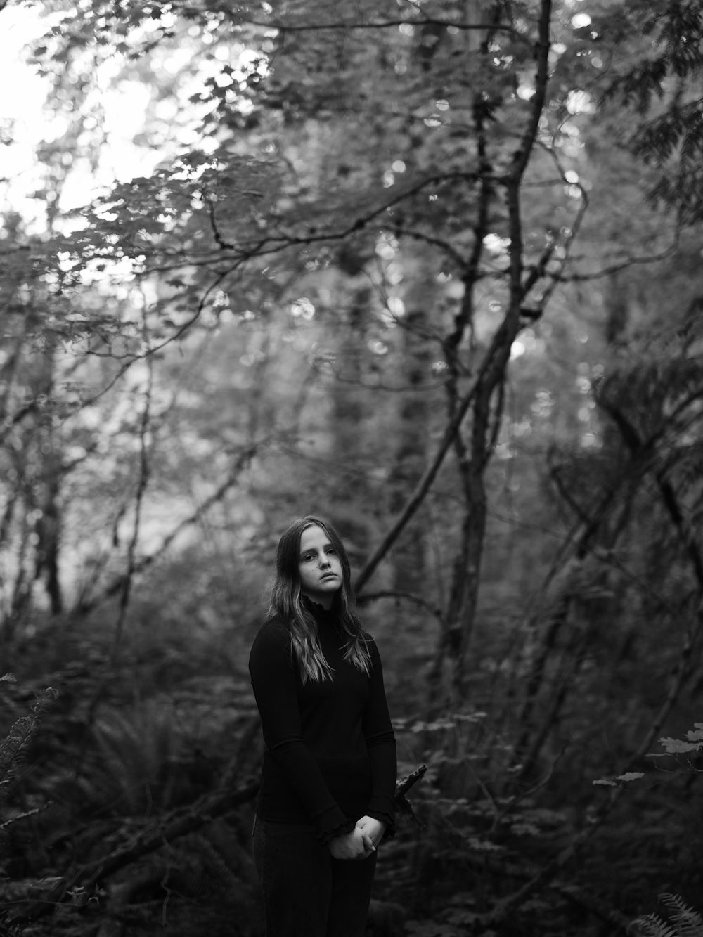 A dark-clad figure stands contemplatively in a misty forest clearing.